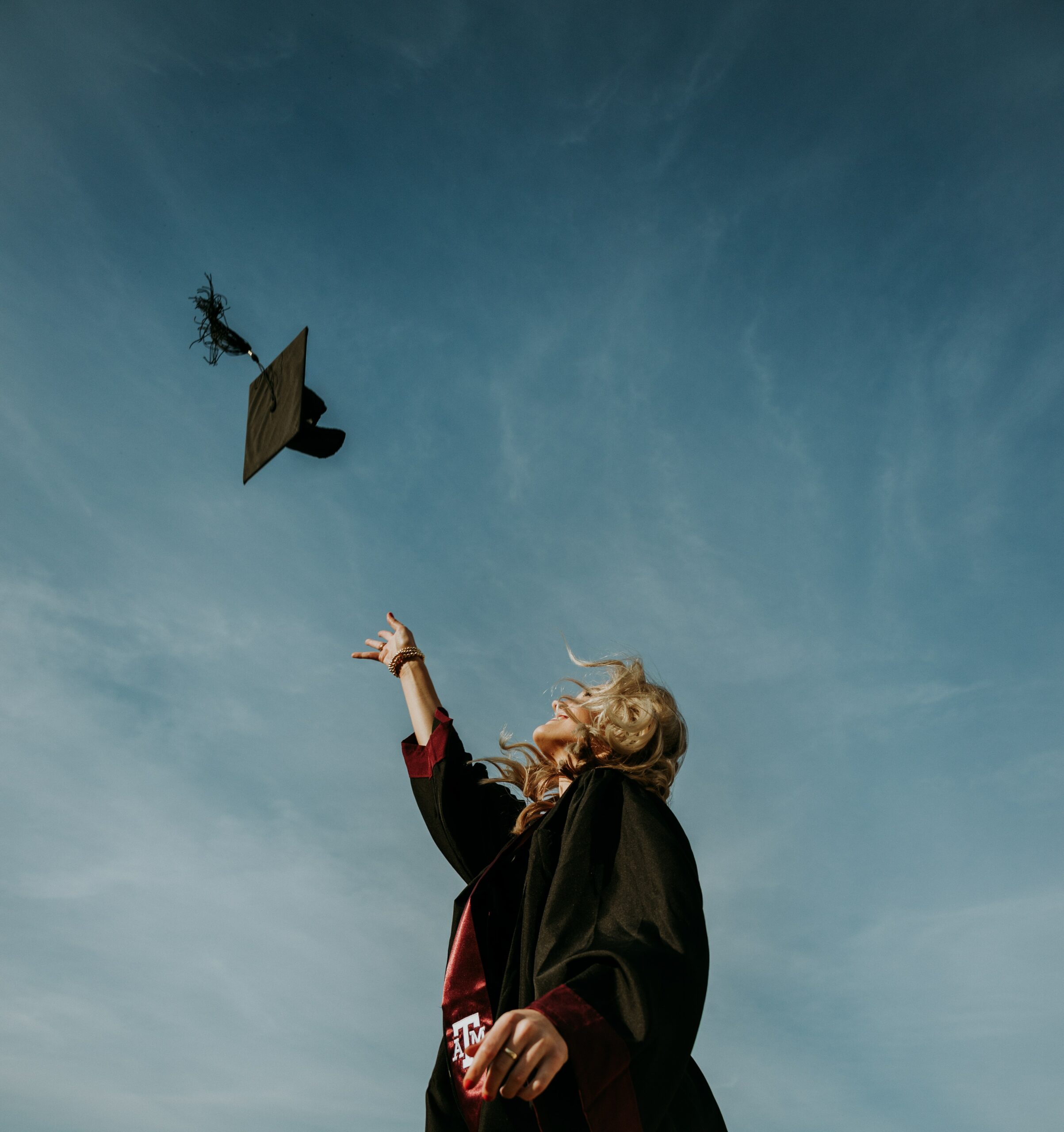 Image of a blond haired female college graduate throwing a graduation cap into the air against a blue sky