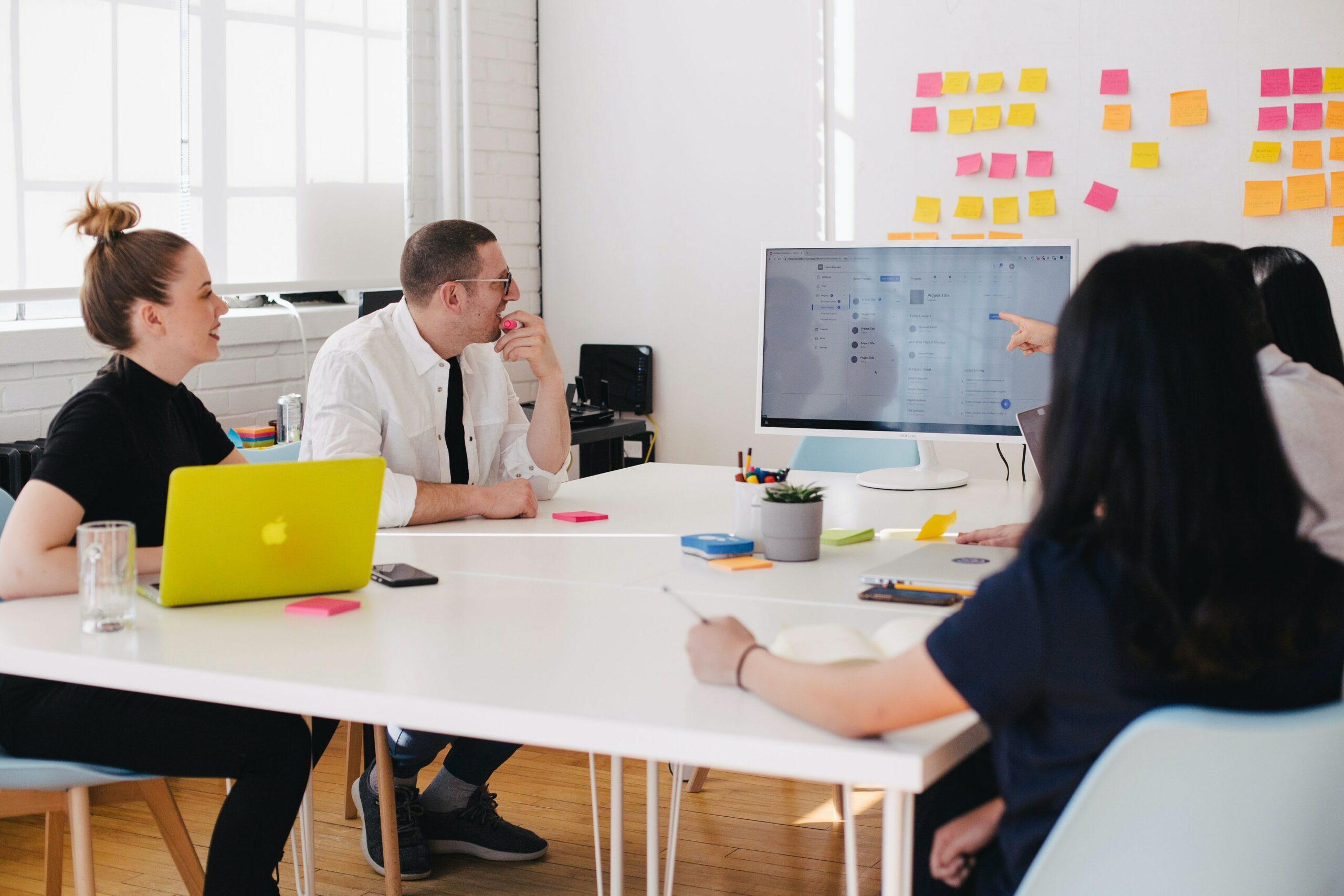 Image of four business people sitting around a white table and looking at a large computer screen with files on it and sticky notes on a wall behind the screen