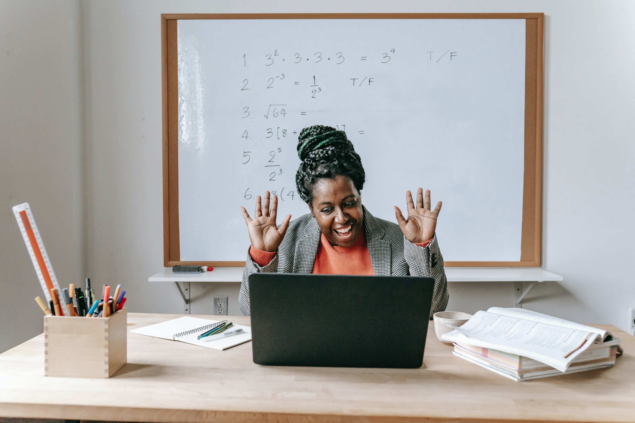 Image of a happy Black woman with a orange shirt and grey blazer using a laptop for online work in a classroom