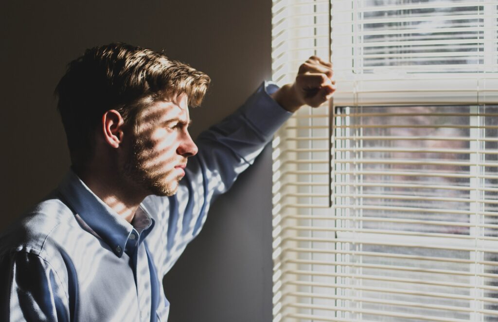 sad man looking out of a window covered in bllinds