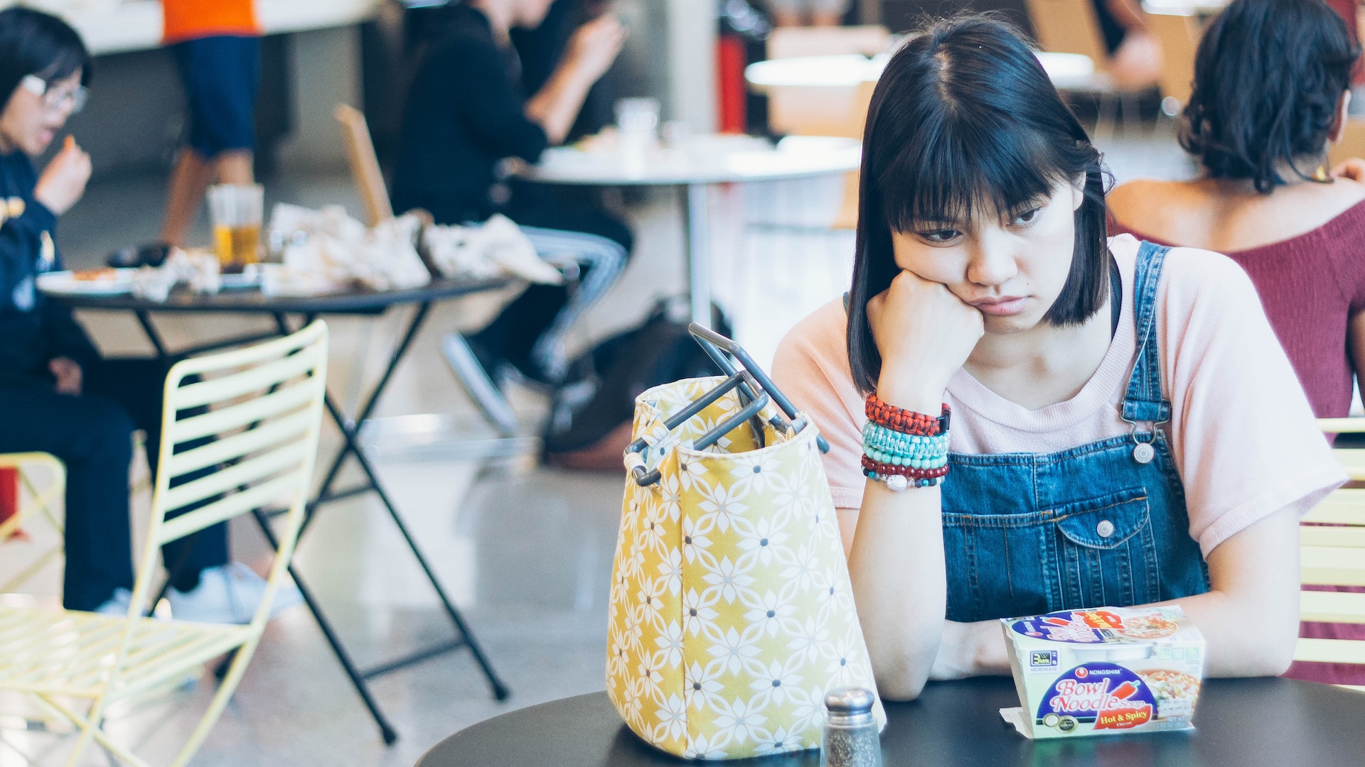 Sad girl with dark hair and overalls eating lunch in a cafeteria